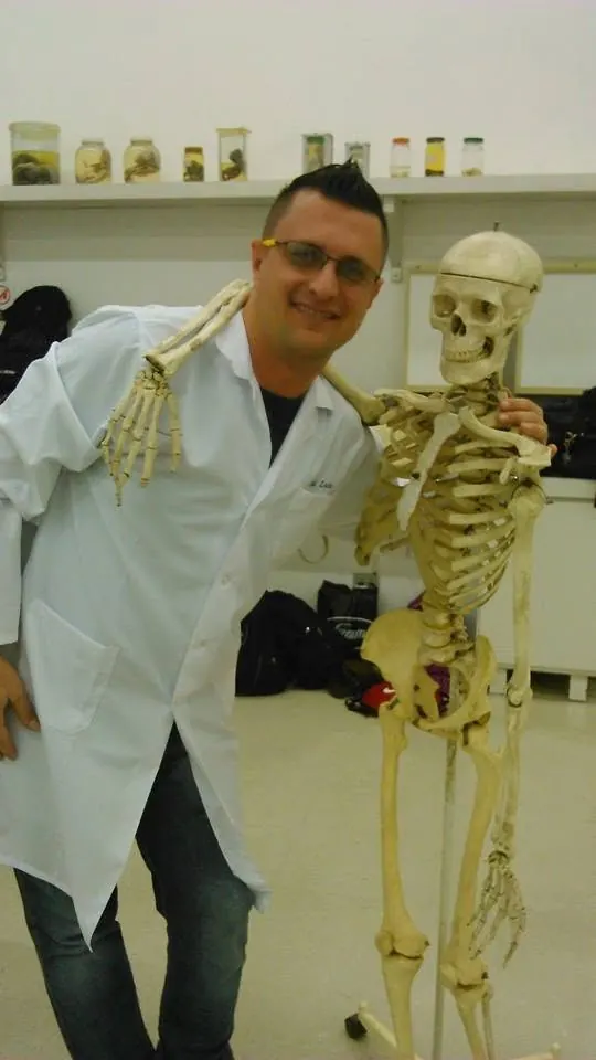 André Luís de jaleco branco, ao lado de um esqueleto, no laboratório da faculdade